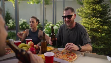 Over-the-shoulder-a-happy-brunette-guy-in-sunglasses-clinks-a-brown-bottle-of-beer-with-his-friend-during-lunch-in-the-courtyard-of-a-country-house