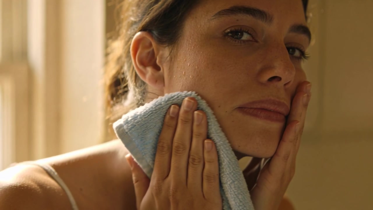 Dabbing woman in camisole drying face and neck at bathroom sink with blue towel after cleansing