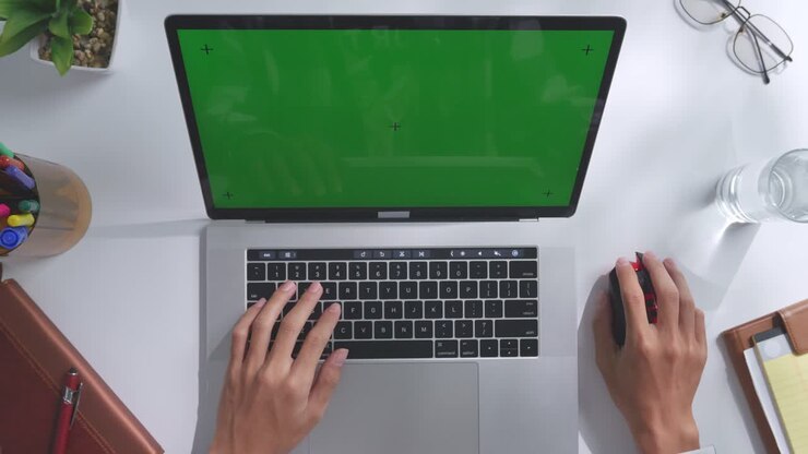 Draufsicht auf einen Laptop-Computer mit Mockup-Greenscreen-Display auf einem Holzschreibtisch, daneben ein Notizbuch mit Stiften, Brille und einem Glas Wasser. Langsames Herauszoomen, Nahaufnahme.