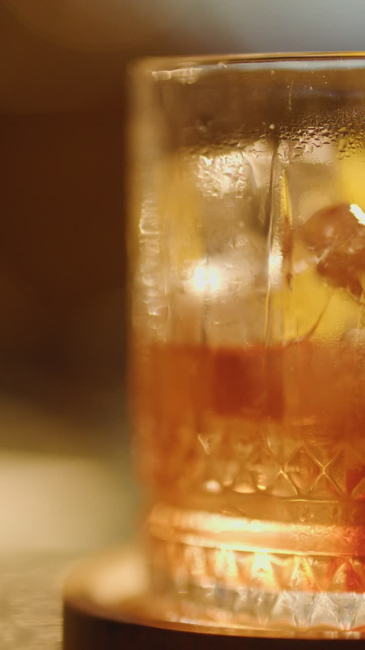 Una sequenza in primo piano mostra una mano che si allunga per sollevare un bicchiere di cocktail Negroni ghiacciato da un bancone del bar sotto un'illuminazione calda e soffusa.