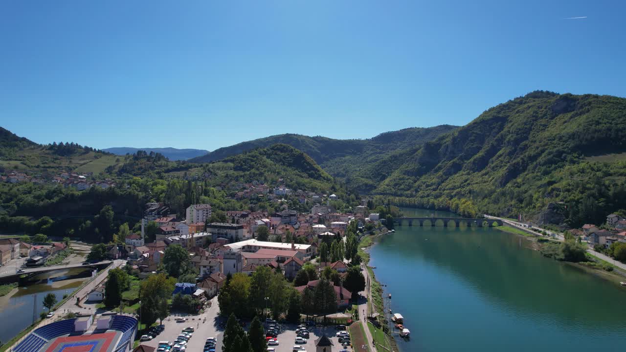 Aerial view of Visegrad in Bosnia and Herzegovina with the Drina river, hillside houses, green mountains, historic stone bridge, riverside roads, boats, and town buildings under clear blue sky