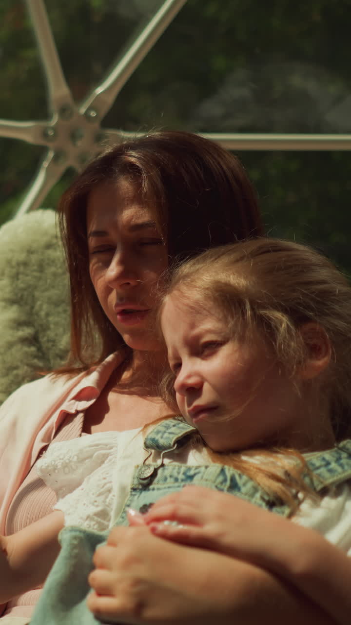 Girl with blonde hair closes eyes to shield from sunlight on transparent terrace. Woman looks out of window with little daughter sitting in soft armchair