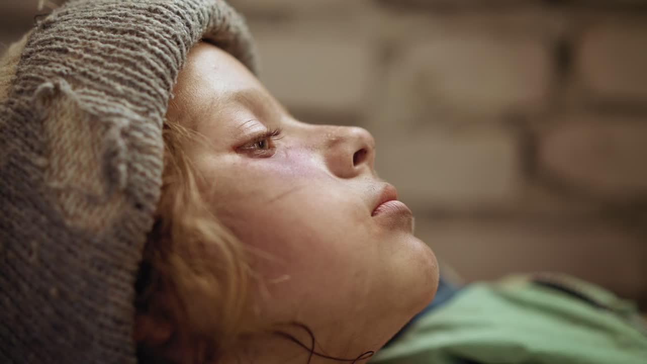 Close up of sick orphan coughing while lying down, cheeks bruised and face dirt streaked, wearing tattered wool hat, eyes half closed in fatigue