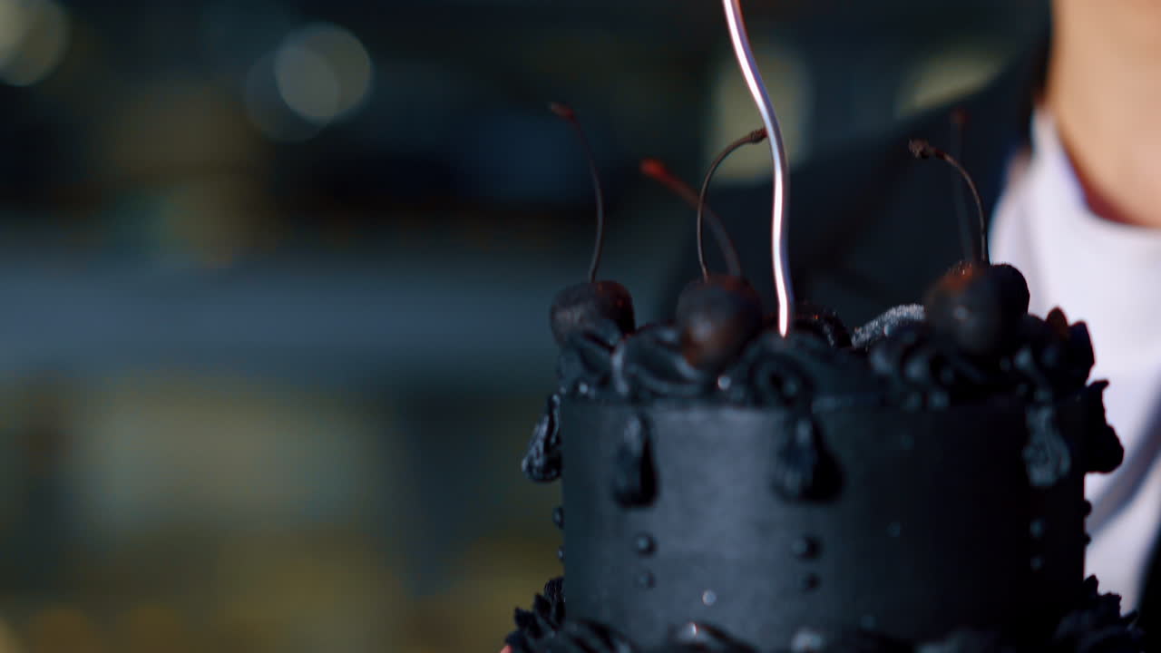 Female hand lighting a thin silver candle on the black cake with cherries. Close up. Blurred backdrop.