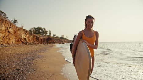 A-blonde-girl-in-an-orange-swimsuit-carries-a-light-colored-wooden-surfboard-and-walks-along-the-sandy-seashore-in-the-morning-at-Sunrise.-A-blonde-girl-surfing-master-walks-along-the-sea-with-her-surfboard