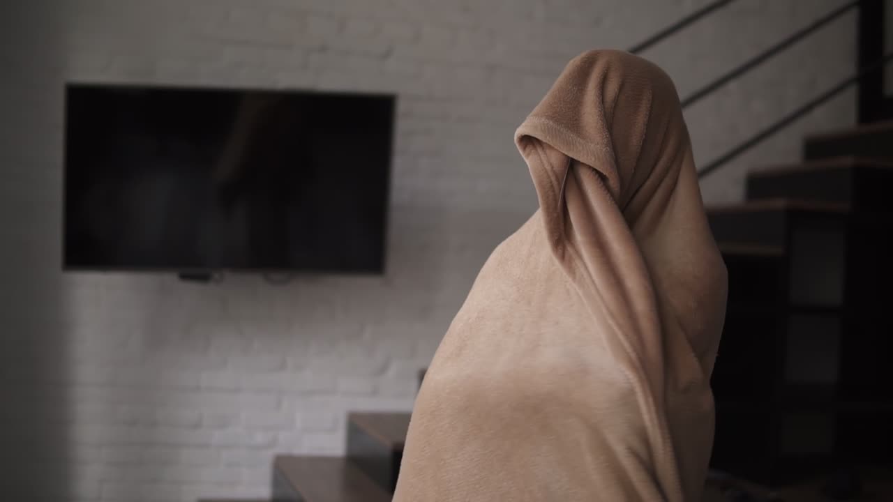 즐거운 여자가 플레이드에 싸여 서서, 미소 짓고 집의 창문을 쳐다본다.