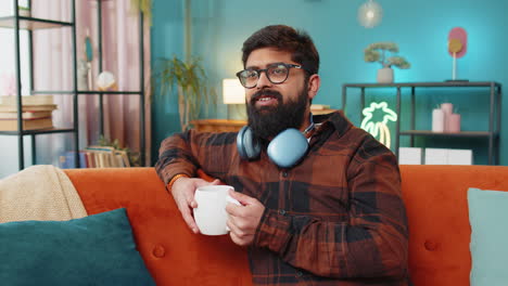 Attractive-smiling-Indian-young-man-drinking-a-cup-of-coffee-or-herbal-tea-at-home-couch-in-morning