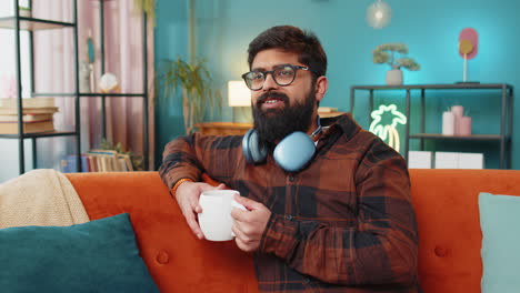 attractive smiling indian young man drinking a cup of coffee or herbal tea at home couch in morning