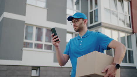 a male mail carrier with round glasses carries boxes for delivery to customers and looks at his mobile phone. search through the mobile phone for the delivery address. payment terminal.