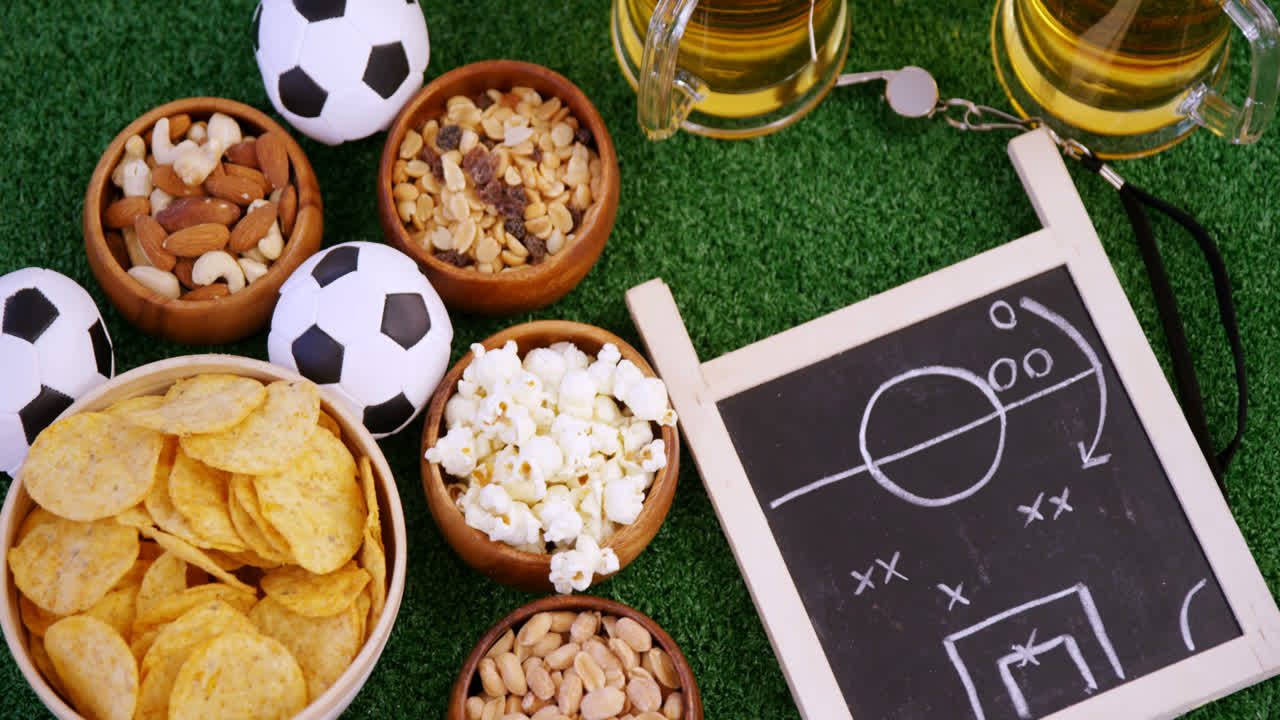Schüssel mit Snacks, Fußball, Bier, Trillerpfeife und Schiefertafel auf Kunstrasen 4k