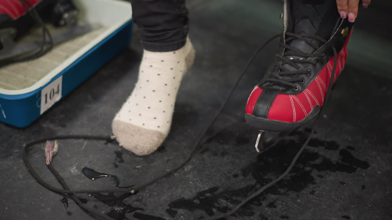 Closeup of red black ice skates placed in blue tray on locker room floor with female wearing polka dot socks and black pants. As she is wearing the ice skates shoe