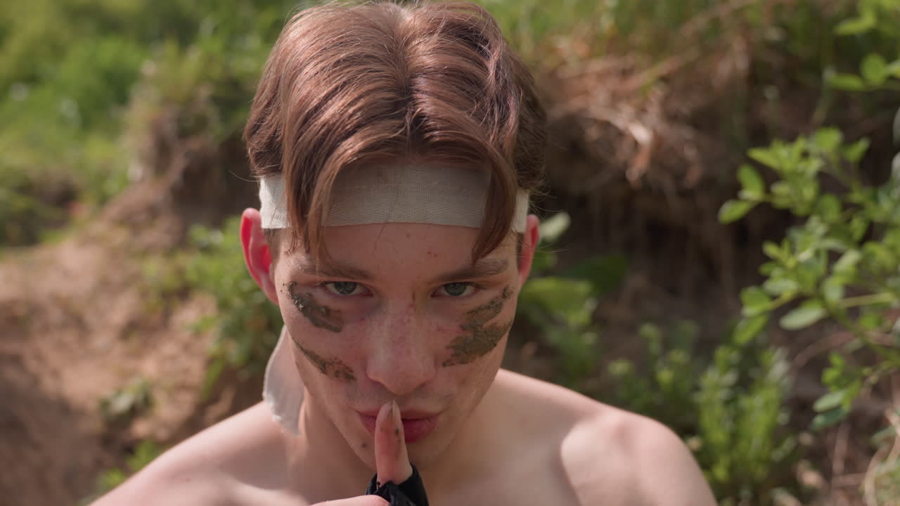 Brown hair tourist stares into camera under bright sunlight, mud paint smeared on cheeks, headband in place. Slow motion reveals calm yet bold expression, conveying silent command on rugged terrain