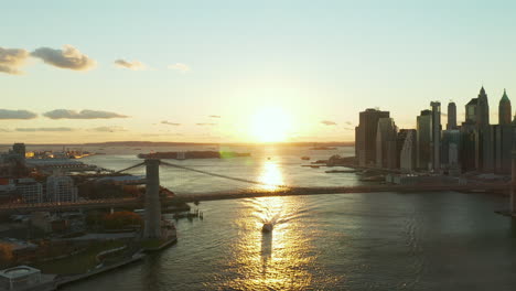 Aerial-panoramic-view-of-Brooklyn-Bridge-against-sunset.-Cruise-ship-floating-of-glowing-water-surface.-Skyscrapers-on-waterfront.-Manhattan,-New-York-City,-USA