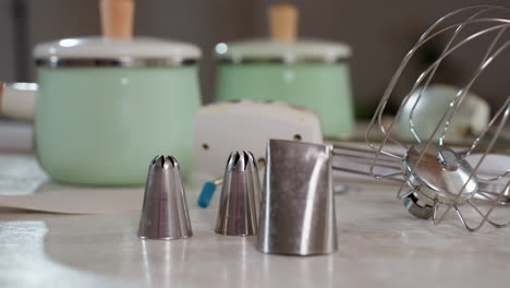 close up of stainless steel piping nozzles and electric whisk attachment on kitchen counter with pastel green ceramic canisters and white appliance in background