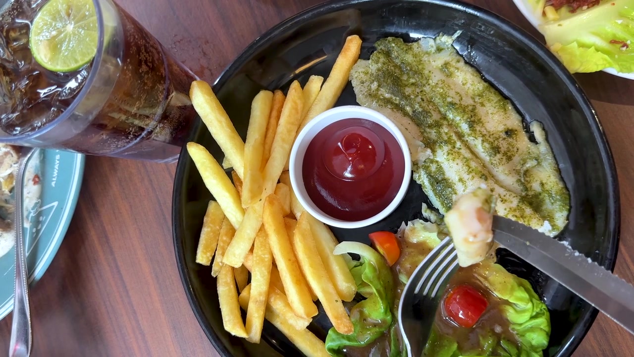 Scatto dall'alto di mani che usano le posate per mangiare pesce alla griglia con patatine fritte e insalata.