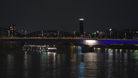 Embárquese-En-Un-Fascinante-Viaje-Nocturno-A-Lo-Largo-Del-Río-Rin,-Mientras-Un-Crucero-Se-Desliza-Con-Gracia-Por-Los-Puentes-Iluminados-De-Colonia,-Creando-Un-Espectáculo-Cautivador.