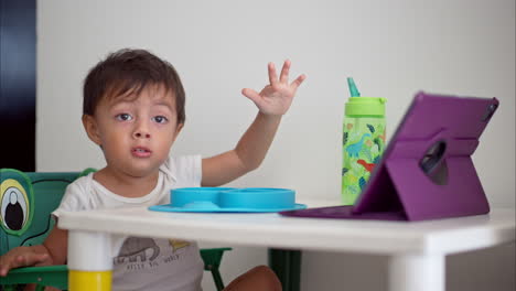 Lindo-Bebé-Latino-Divirtiéndose-Viendo-Una-Película-En-Una-Tableta-Morada-Mientras-Desayuna-Sentado-En-Una-Pequeña-Mesa-Blanca-Para-Niños