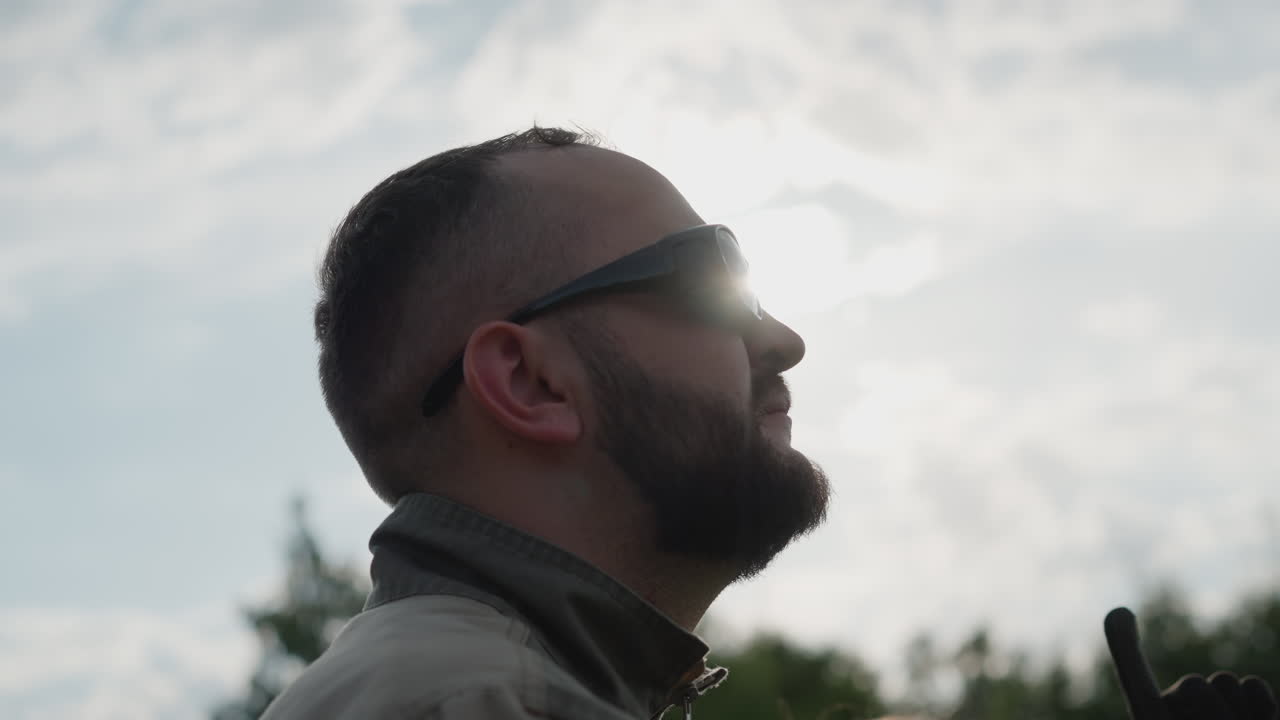 close up young man wearing dark sunglasses gazing upward toward bright sky, sunlight halo behind head, subtle lens flare and blurred tree line backdrop