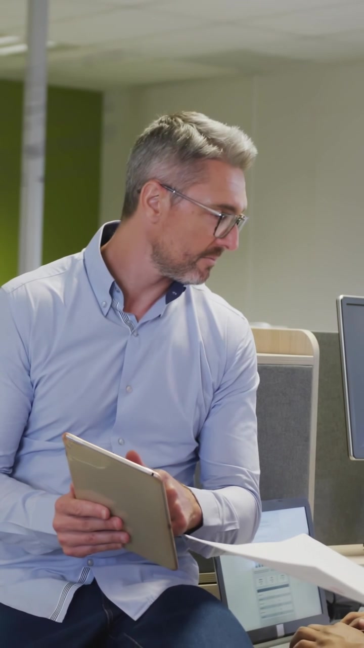 Video verticale: Il leader che prende pagine dal collega, indicando un tablet e uno schermo per una revisione aziendale.