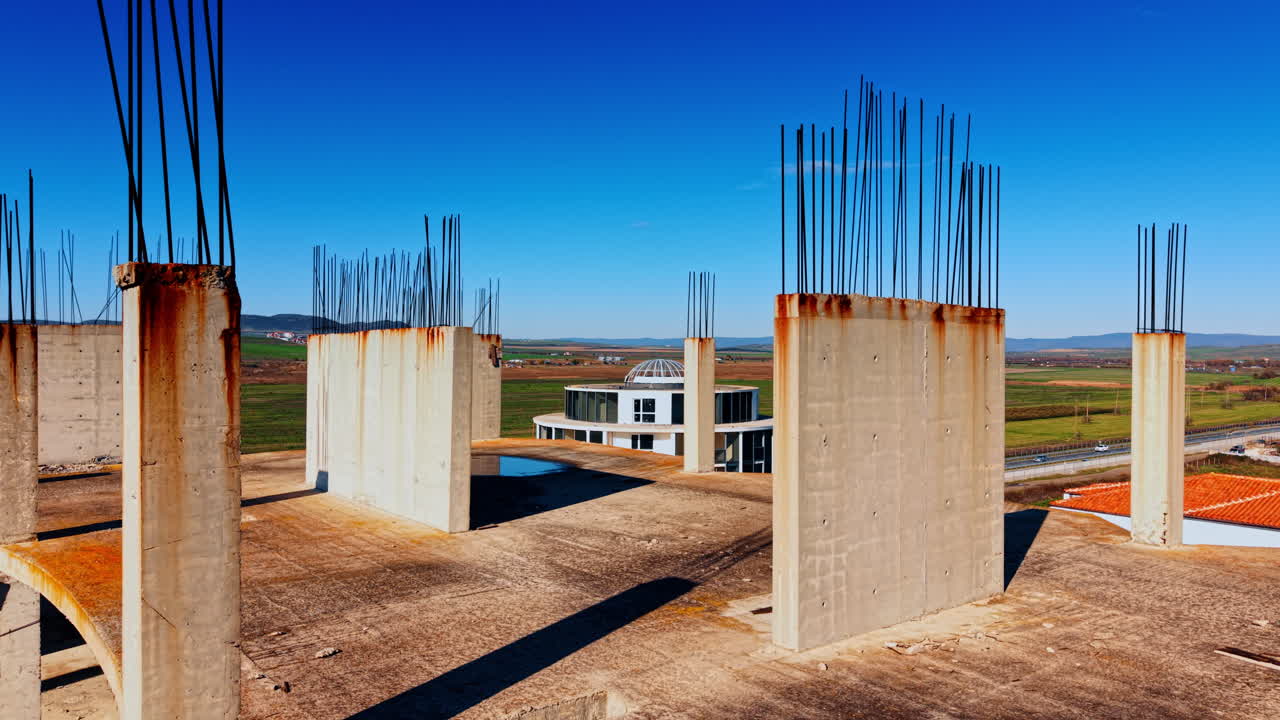 Colonne e solai con tondini metallici sporgenti si trovano in cima alla costruzione incompiuta. Vasti campi verdi, catena montuosa e cielo blu sullo sfondo