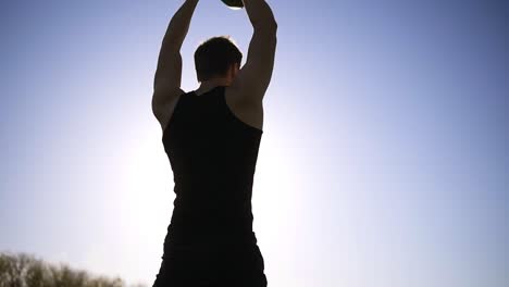 a man, with a beautiful body sculpture, trains with weight. outdoors. blue sky. backside view