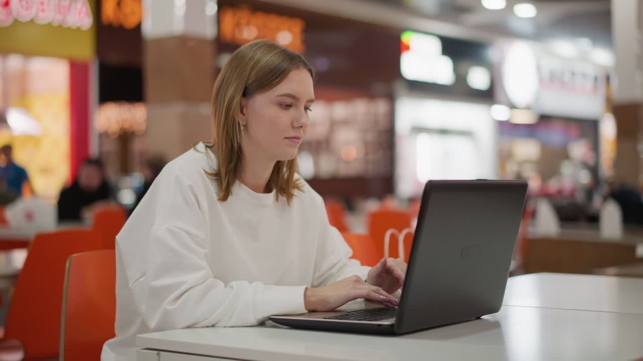 mujer joven enfocada trabajando remotamente en un vibrante centro comercial sentada en una mesa escribiendo en una computadora portátil, atmósfera casual y moderna con fondo borroso con luces de tiendas coloridas y actividad