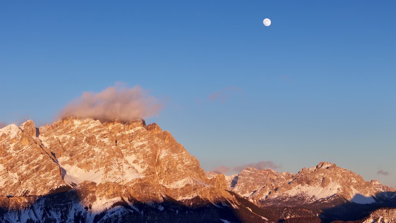 Golden hour in Dolomites with the sun shining on the mountains and the moon right above them (drone footage)