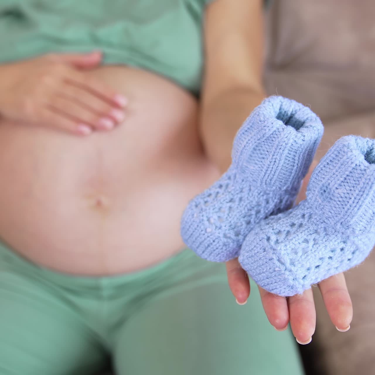 Expectant mother holding pretty blue baby socks on her palm. Mother caressing her big belly lovingly. Knitted tiny socks for newborn boy close up
