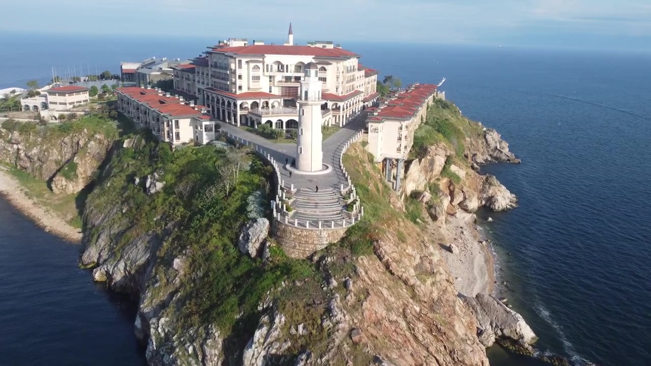 Maestoso video con drone mostra un imponente complesso costiero con tetti rossi e un alto faro bianco su scogliere scoscese sopra il profondo Mar Baltico blu, bagnato dalla chiara luce del sole mattutino