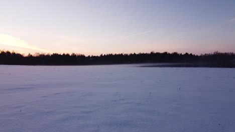 Beautiful-low-angle-aerial-flight-above-snowy,-frosty-field,-misty,-foggy-evening-landscape-after-the-sunset,-wide-angle-drone-shot-moving-forward-low-to-the-ground