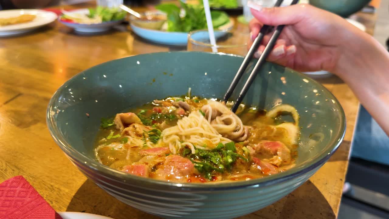 Chopsticks lift rice noodles from a steaming bowl of beef pho in a vibrant Bangkok restaurant setting