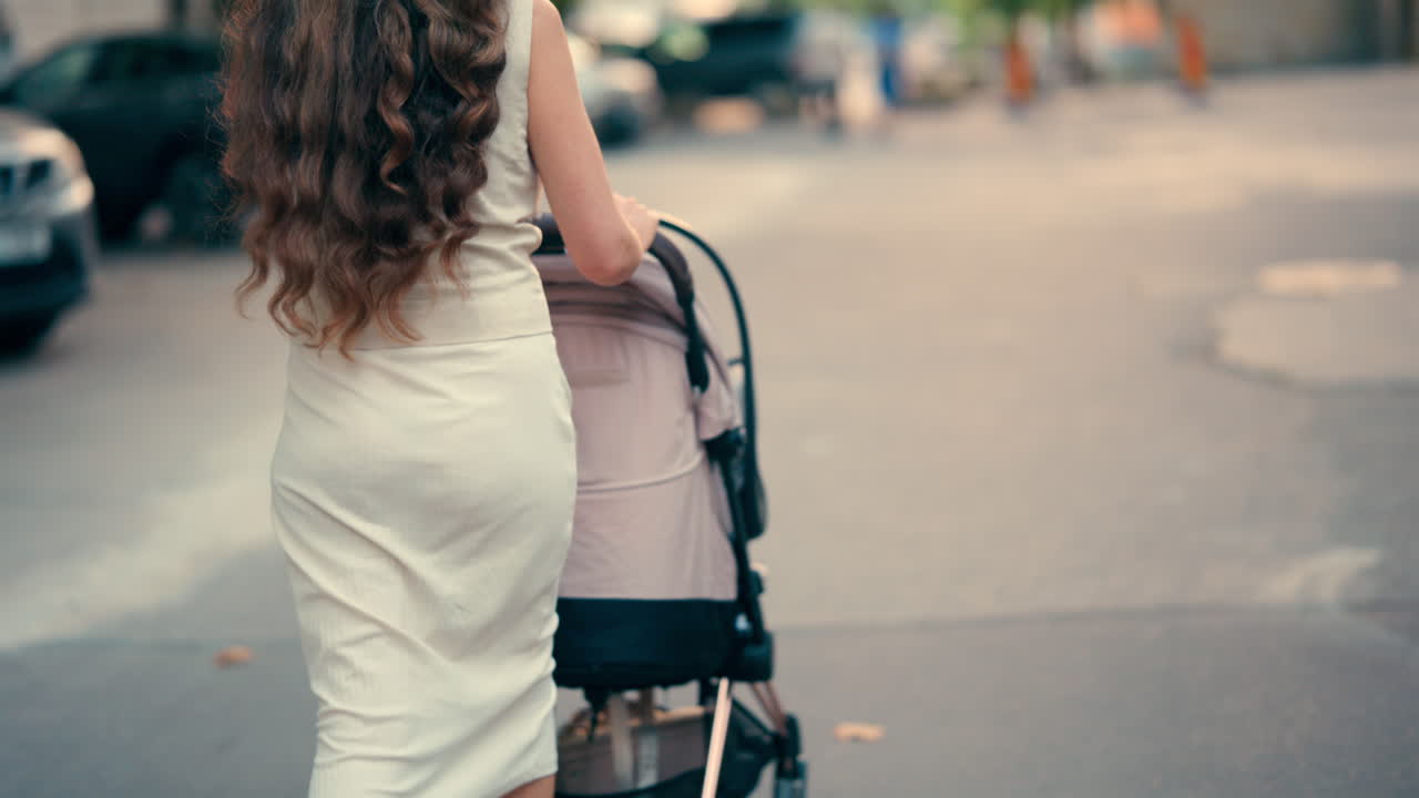 View from the back of a woman with long wavy hair pushing a stroller along a tree-lined park pathway