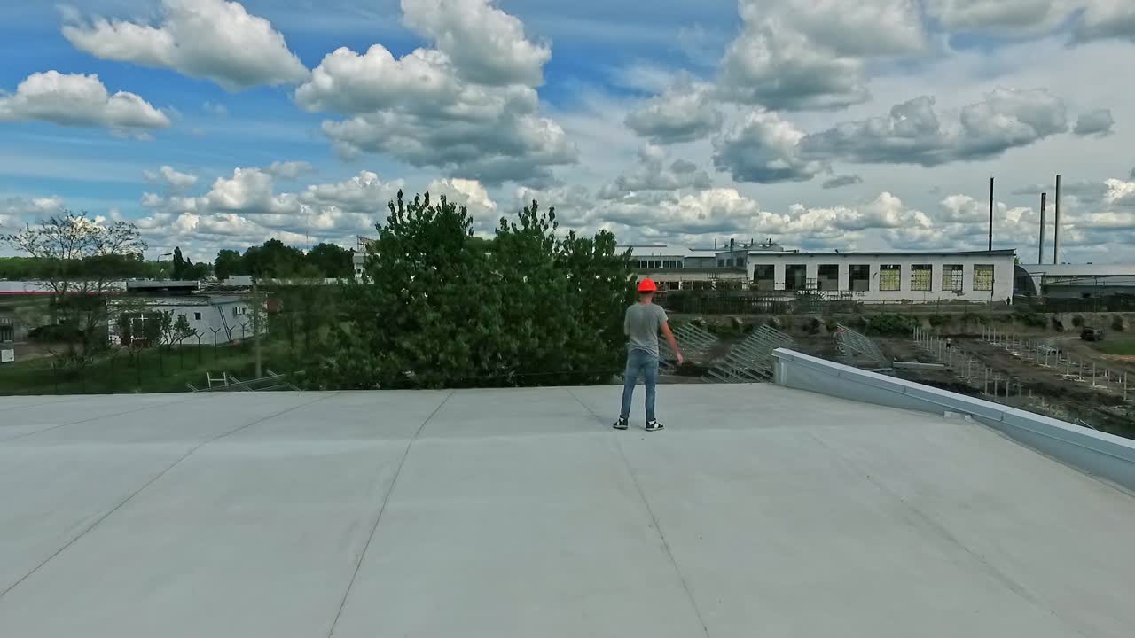 Engineer expert walking on roof. Worker in orange helmet think about new solar farm construction on rooftop of a building on nature background. Aerial view. Motion camera around.