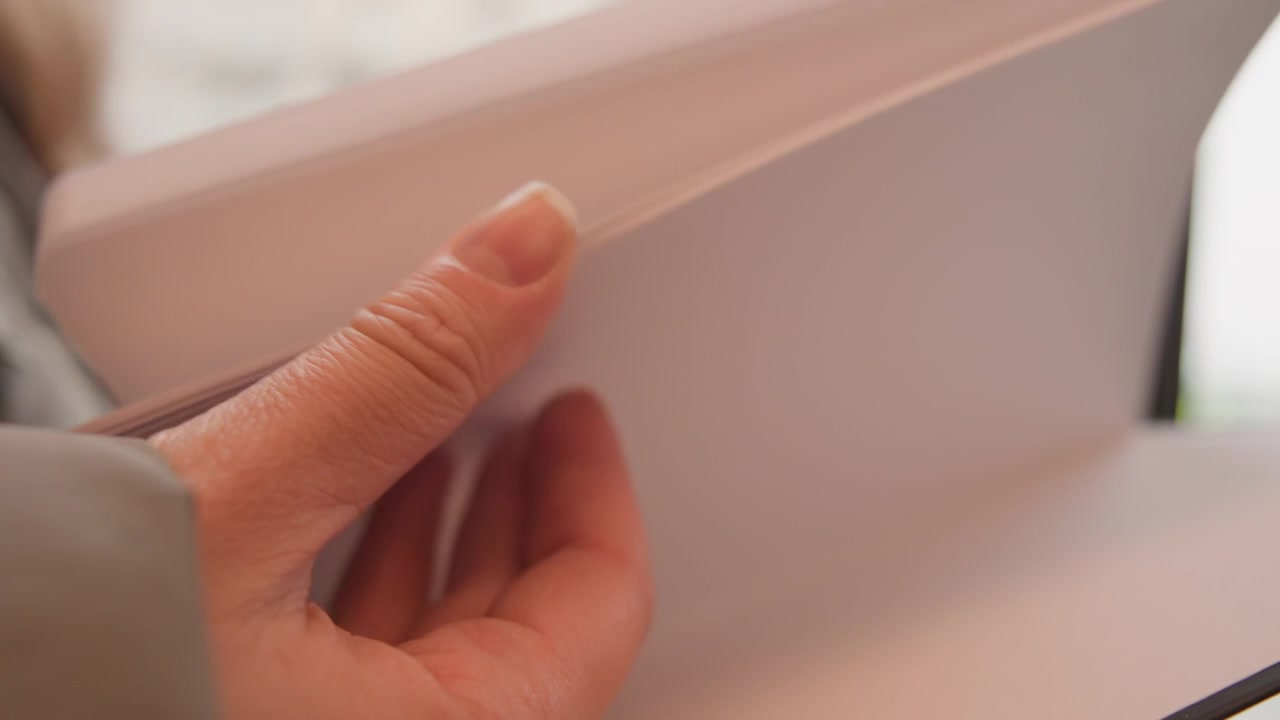 Primer plano de manos pasando páginas en blanco en un cuaderno, luz suave de ventana y fondo de ciudad borroso, ambiente acogedor de cafetería, persona previsualiza maqueta de boceto, estudiante revisa apuntes, escritor planifica diseño de cubierta beige.