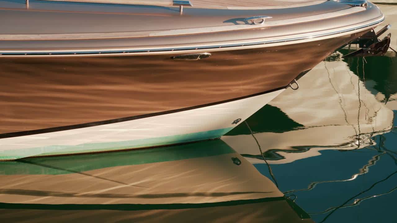 Close up of a yacht's bow shining under warm sunlight, its sleek shape mirrored beautifully on the still green water