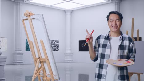 아시아 남성 예술가 가 갤러리에서 오일 페인트와 브러쉬로 그림을 그리면서 캔버스를 가리키고 있다