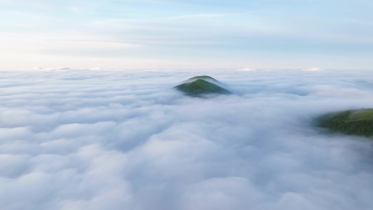 Elevated aerial view of a solitary green hill peak emerging above a dense blanket of low clouds at dawn. Soft morning light and cool blue tones highlight the
