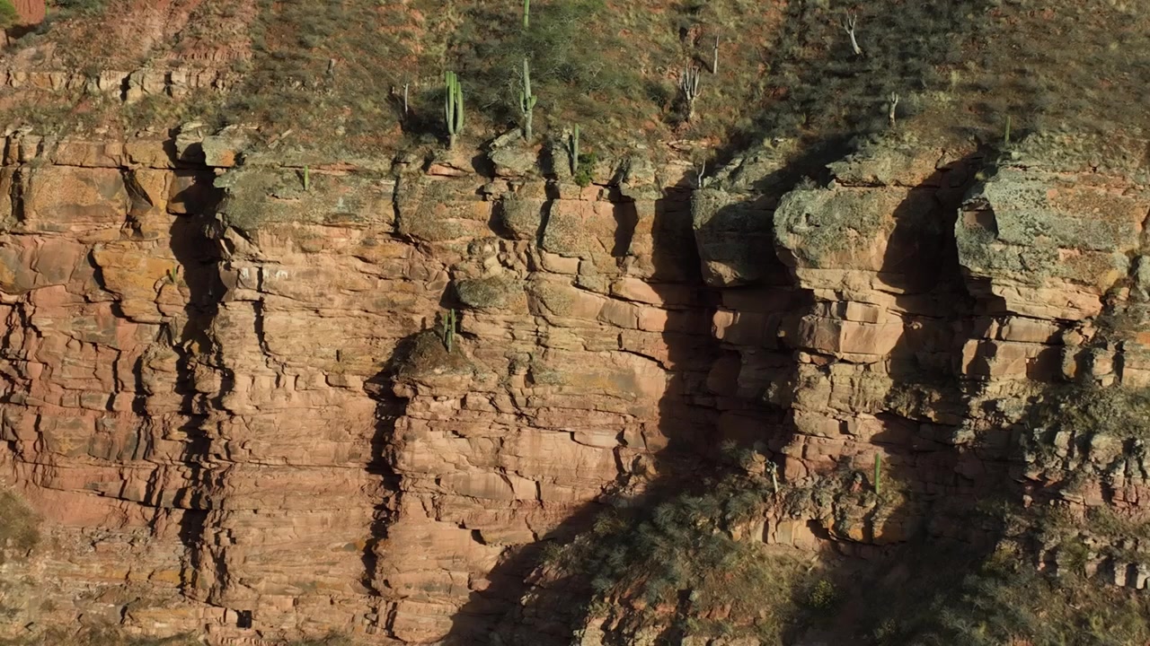 Um drone voa suavemente sobre as montanhas em Salta, Argentina. Cactos característicos da região norte da Argentina são visíveis. O corte na montanha revela a geologia local
