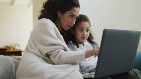 madre e hija de raza mixta felices sentadas en el sofá, divirtiéndose y usando la computadora portátil