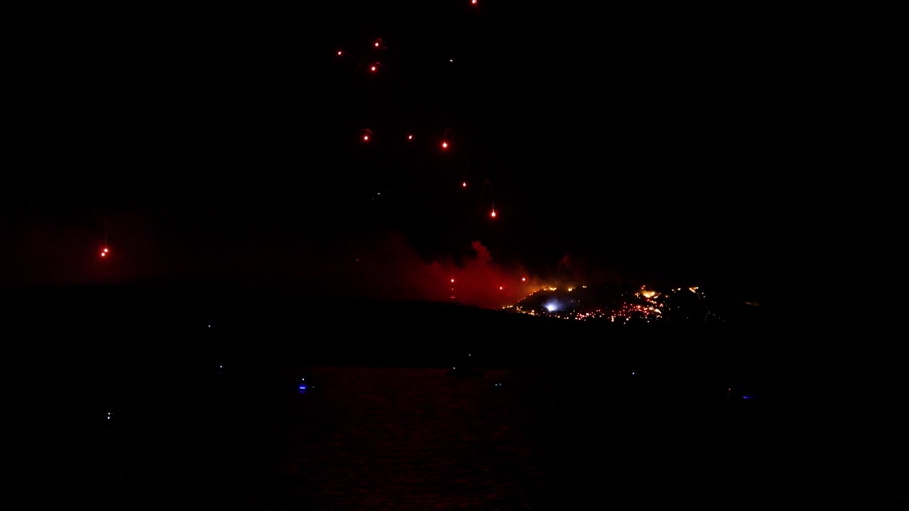 Spectacular night fireworks simulate a volcanic eruption at the Ifestia festival, illuminating the skyline with glowing bursts and smoke over a dark landscape in slow motion.