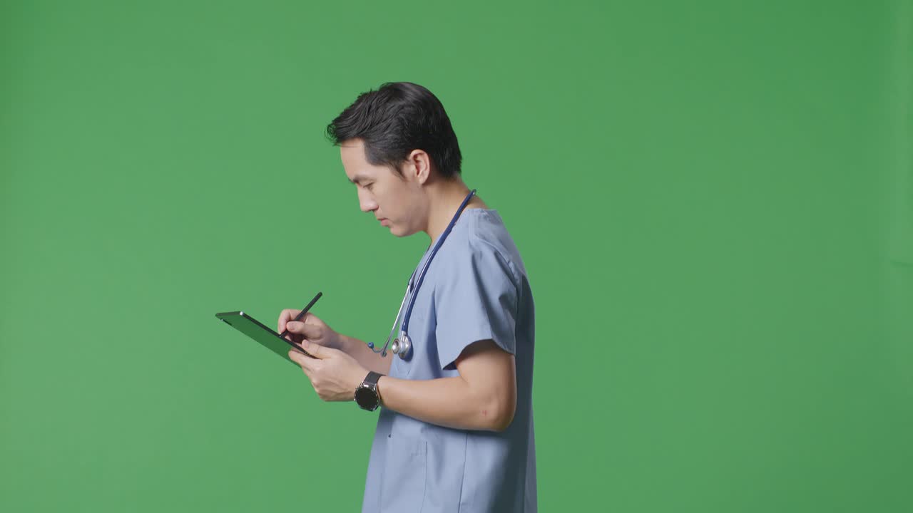 병원 에서 녹색 스크린 배경 에 걸어 다니는 동안 스테토스코프 를 들고 태블릿 에 메모 를 하는 아시아인 남성 의사 의 측면 시각