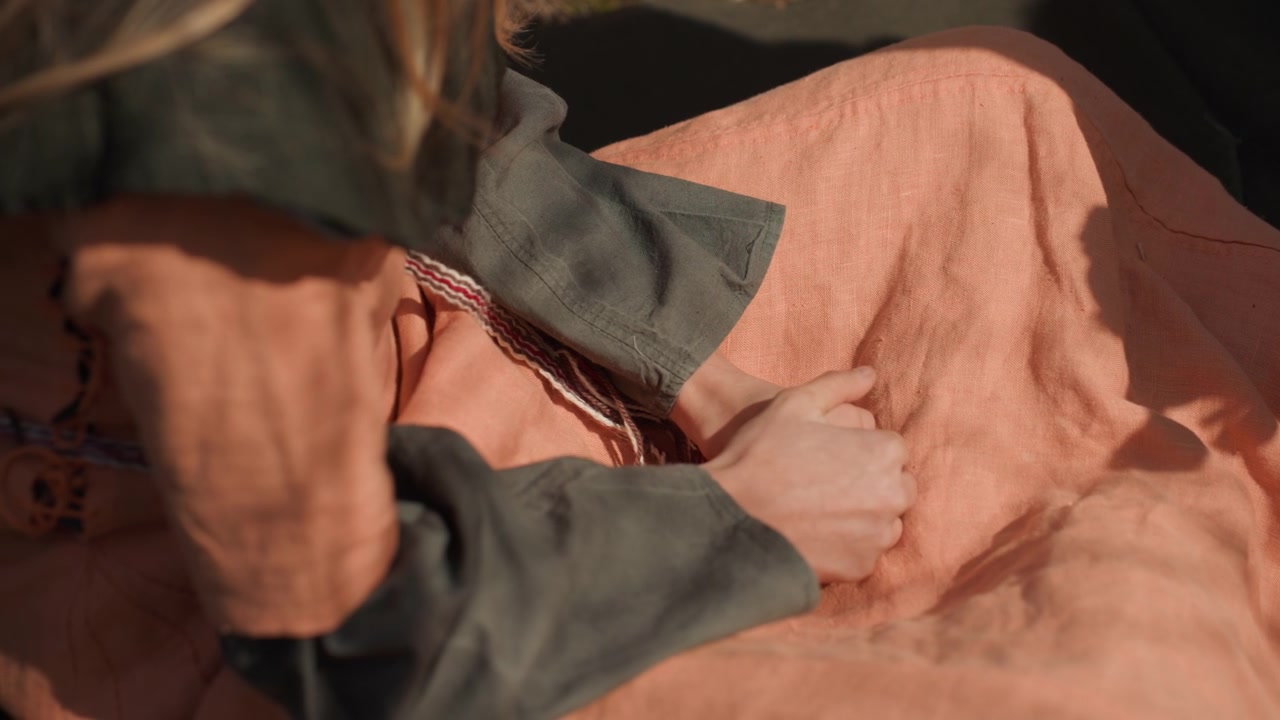 Primo piano ravvicinato dal basso che mostra mani che accarezzano e afferrano una coperta tessuta mentre si è seduti, con un sottile movimento delle dita che preme il tessuto a maglia e sposta le pieghe sotto una calda luce direzionale.