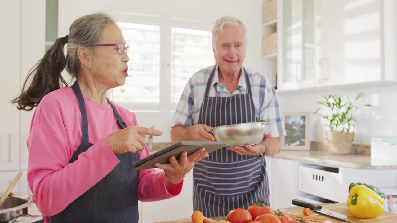 feliz pareja de ancianos diversos usando tableta y cocinar en la cocina