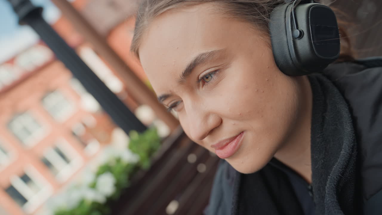 Ung kvinne som nipper til kaffe med hodetelefoner på en solrik terrasse på en kafé i byen, engangskopp mot leppene, et mildt smil og avslappet blikk, oppmerksom lytting til podcast, koselig jakke og mykt omgivelseslys