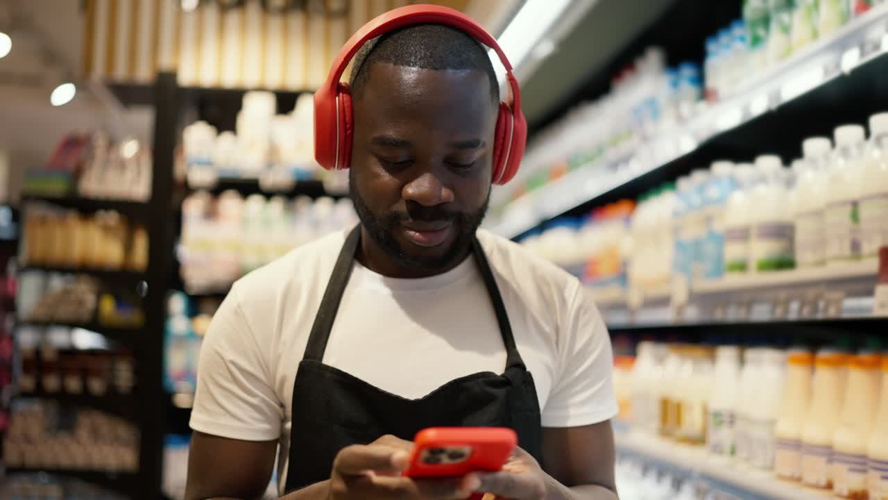 a man with Black skin color in red headphones and a black apron with a red smartphone walks along the dairy department in a supermarket