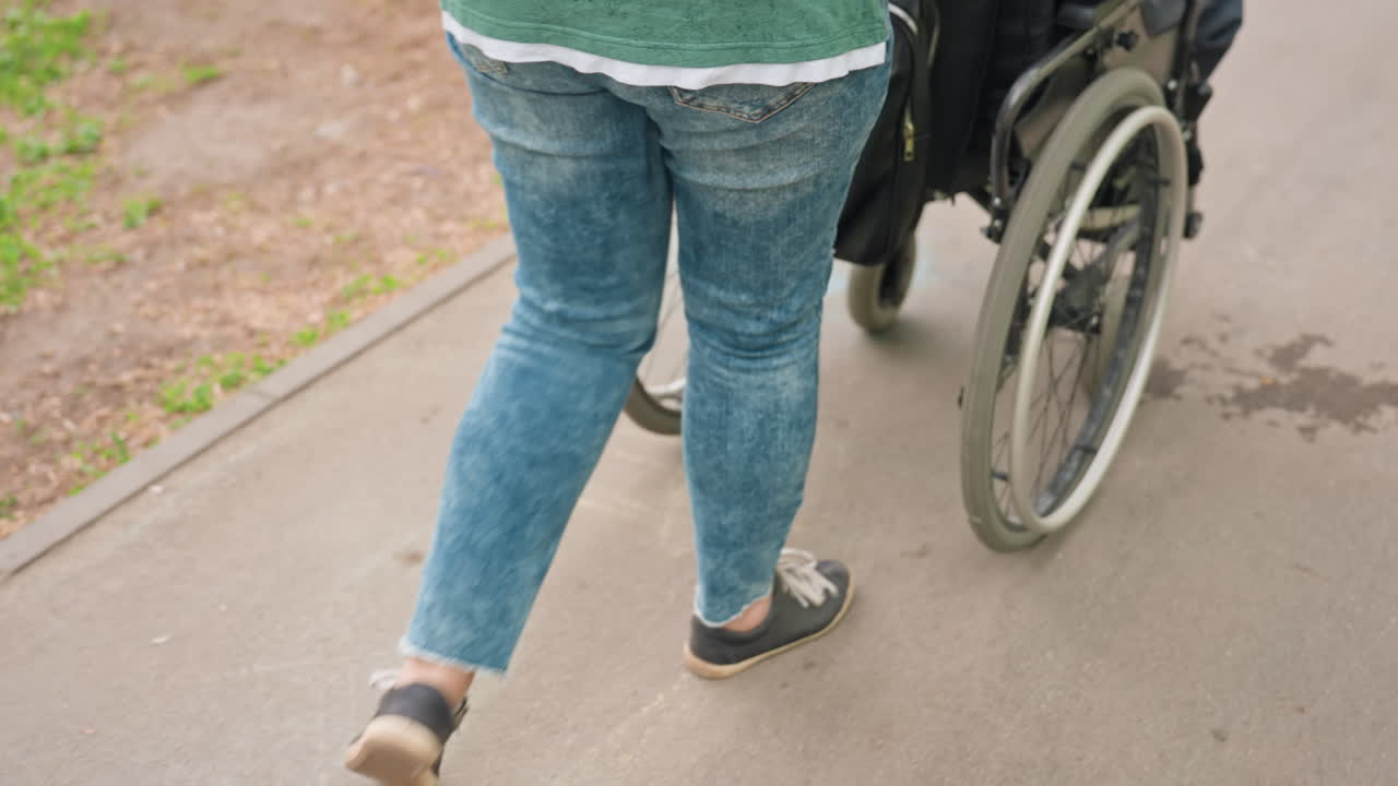 Walking Behind Wheelchair On Park Path, Jeans And Denim Jacket Visible, Stride And Wheel In Frame, Candid Companion Role Showing Steady Pace And Attentive Support In Green Outdoor Setting