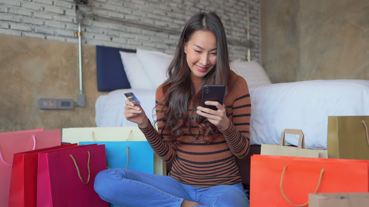 Smiling Woman holding credit card enters numbers on her cellphone, siting bedroom floor surrounded shopping bags