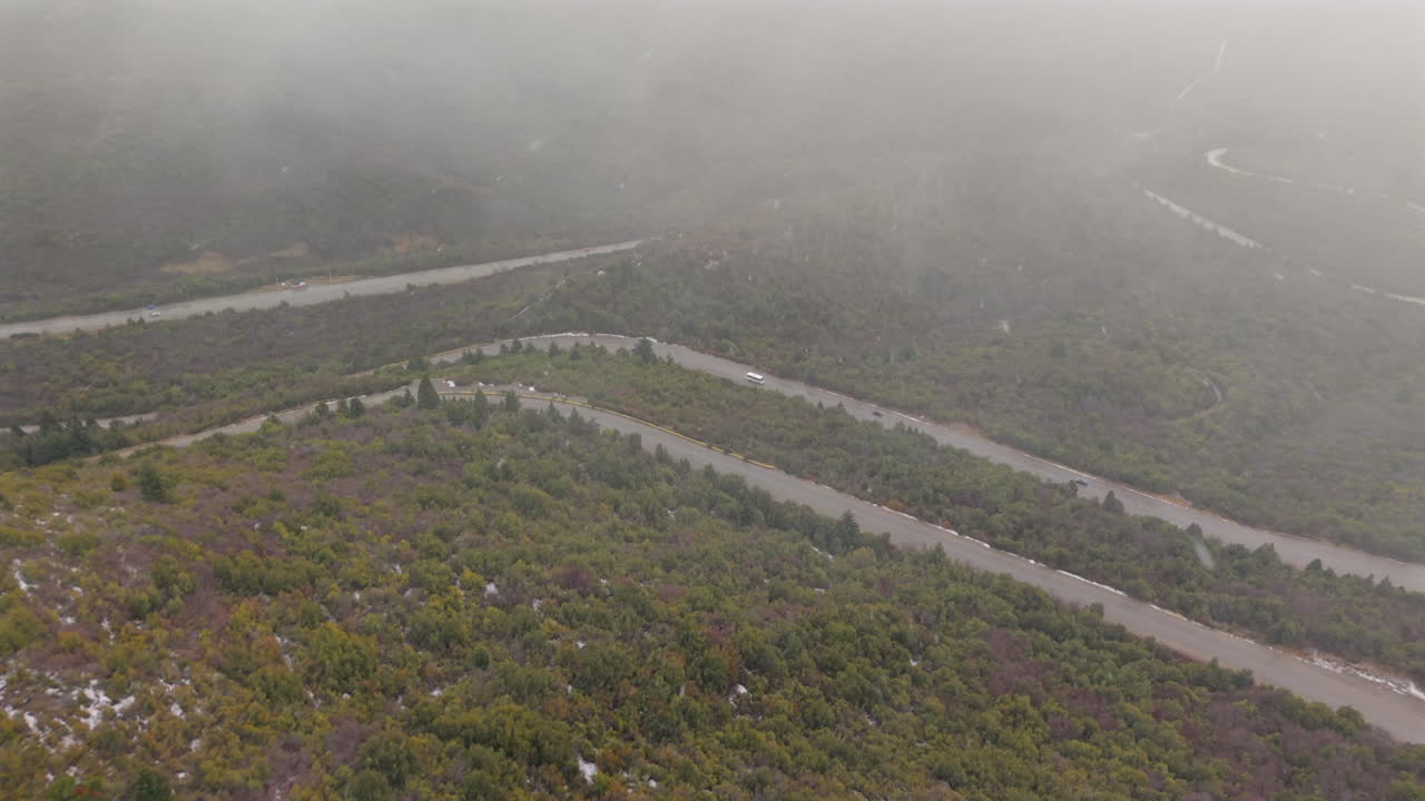 Drone captures winding mountain road carving through Patagonian forest in low visibility as snowstorm sweeps the hills near Bariloche, Argentina—subdued car lights barely visible through swirling mist
