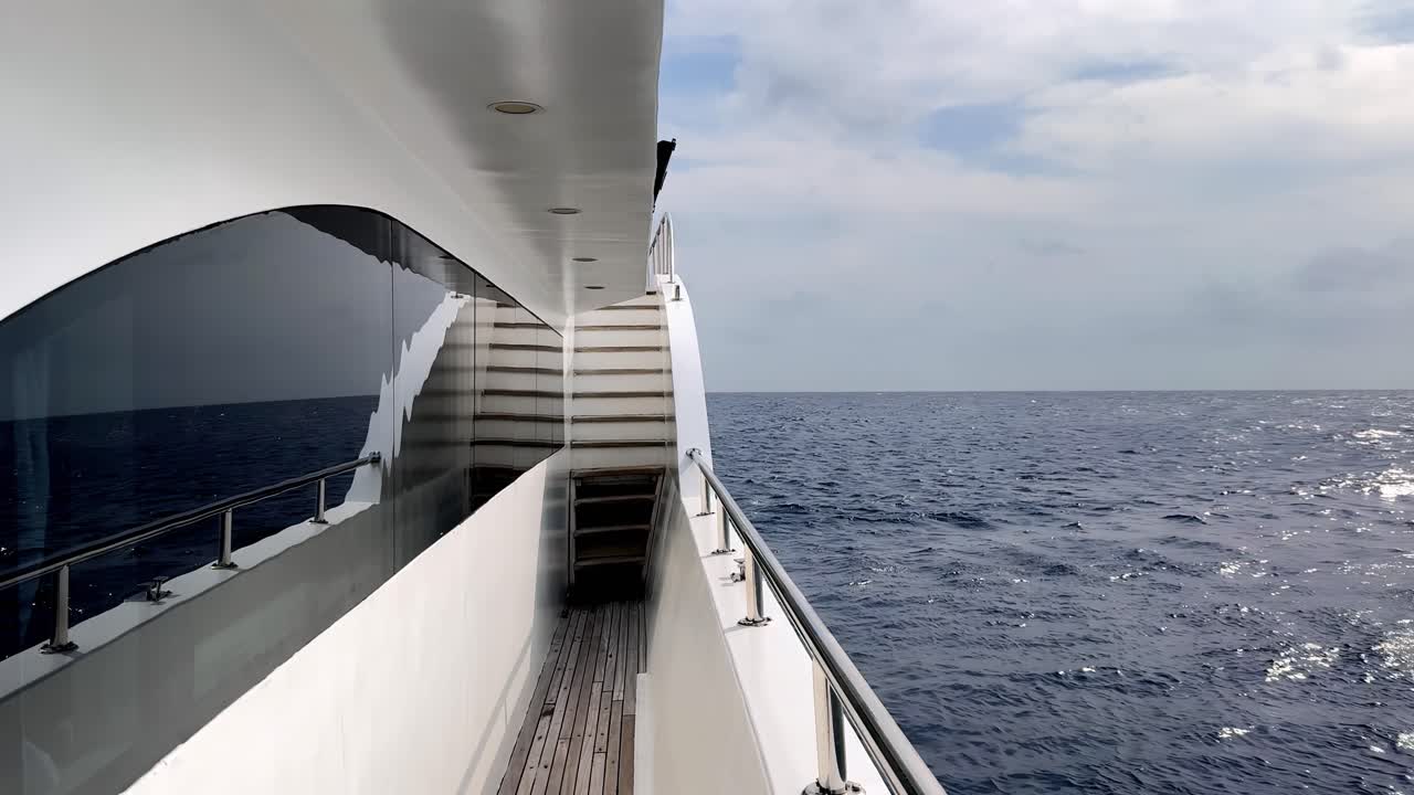 Luxusyachtdeck mit Blick auf das tiefblaue Mittelmeer an einem hellen Sommertag mit sanften weißen Wolken und schimmernden Sonnenlichtreflexionen auf den Wasserwellen, aus einer niedrigen Seitenperspektive