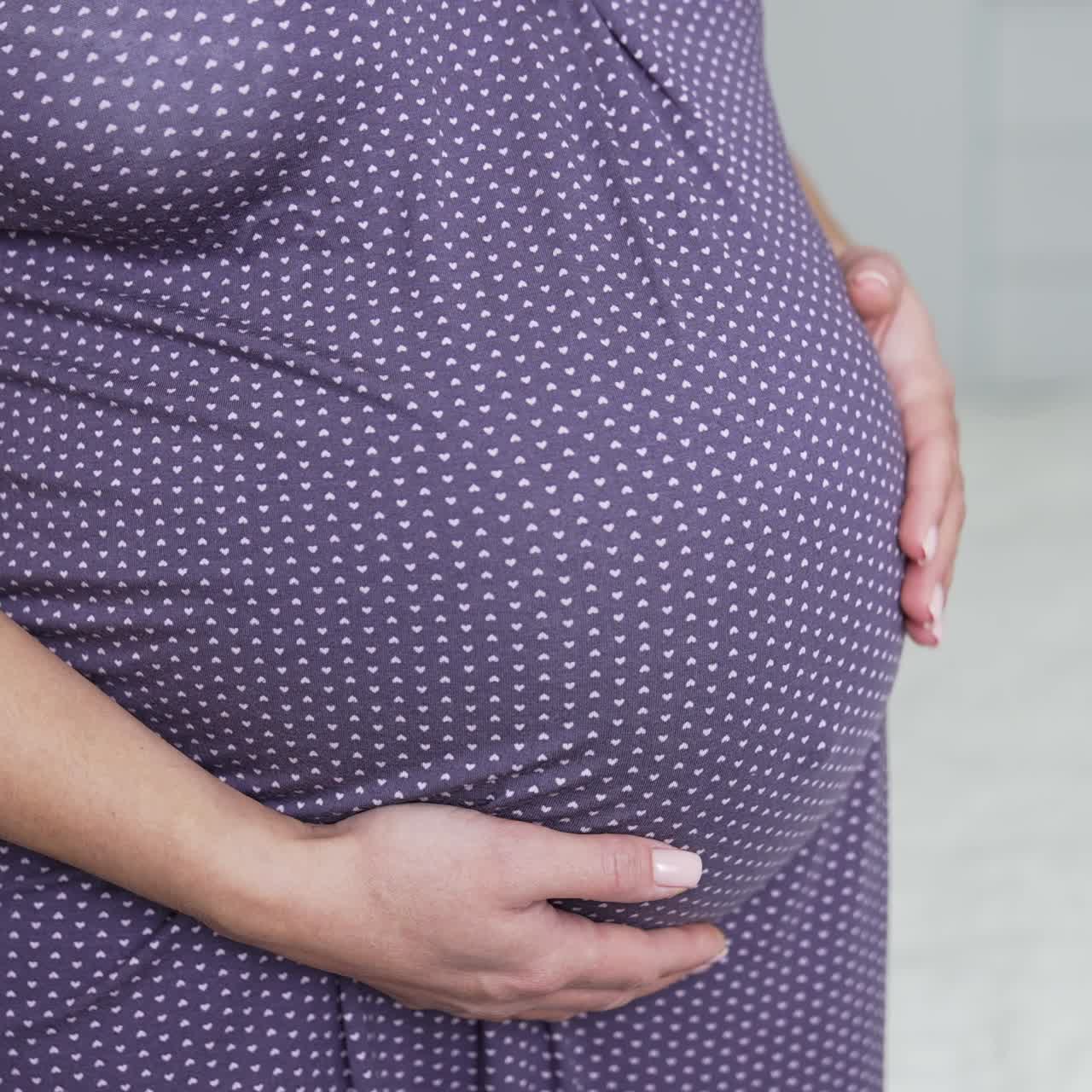 Expectant lady standing half-turned in her bedroom by the bed. Woman expecting a baby wearing a nightgown and stroking her belly. Close up